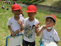 草が生い茂っている河川敷のような所で帽子を被った3人の児童達がポーズをとったり笑顔で微笑んでいる写真