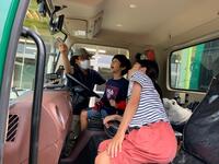 1人の大人が車の説明をしているのを2人の児童が運転席に座りながら興味深そうに聞いている写真