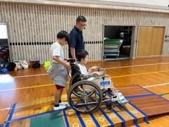 二人の児童達が車椅子に乗る側と押す側に分かれ講師の方に付き添われながら車椅子を押す練習をしている写真