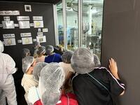 不織布キャプを被った複数の児童達が興味深そうにガラス越しの作業場を見ている写真