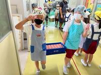 エプロンと三角巾を被った児童2人が牛乳パックが沢山入ったケースを持っている写真
