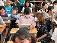 教室のようなところで児童が席についてタブレットを操作している様子を大人がしゃがんで見守っている写真