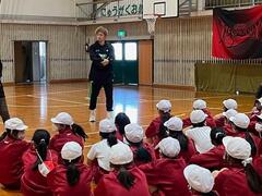 体育館のような所に、男性1人が立っておりその様子を帽子を被った児童達が座って見ている写真
