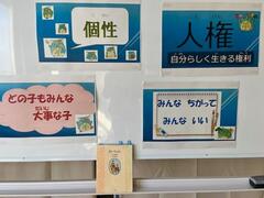 ホワイトボードに「個性」「人権」「どの子もみんな大事な子」「みんなちがってみんないい」と書かれた4枚の紙が貼られており、粉受に本が置かれている写真