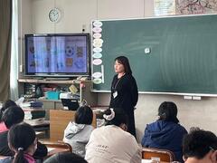 テレビと黒板の手前に1人の女性が立っているのを席に着席した児童達が見ている写真