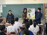 黒板前に、2人の大人がポスターを広げ持っておりその左側に男性が立っている様子を複数の児童達が見ている写真