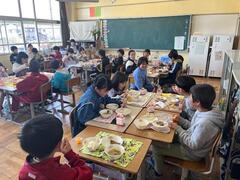 黒板がある教室のような所で複数の児童達が各々机を合わせて給食を食べている写真