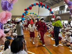 体育館のような所でペーパーフラワーで作られたアーチ状の中を2人の児童が手を繋いで歩いているのを沢山の児童達が座って見ている写真