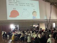体育館のような所の壁にスクリーンが映し出されているのを沢山の児童達が座って見ている写真