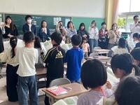 教室内で児童が本を持って立ち、大人達の方を見ながら音読を行っている写真