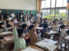 教室のような所で席についた複数の児童達が手を挙げており、その様子を大人達が後ろで見守っている写真