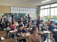 教室のような所で席についた複数の児童が手を挙げたりしているのを複数の大人が見ている写真