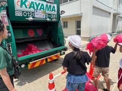 ゴミ収集車の回収口に、ゴミに見立てた袋のようなものを投げている児童達の写真