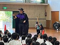 体育館で帽子を被りマントのようなものを身に纏った男性を複数の児童が見ている写真