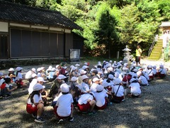 神社の境内のような所で1人の男性が立っており、その様子を帽子を被った沢山の児童達がしゃがんで見ている写真
