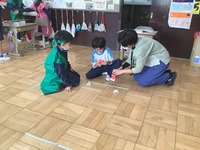 1年生におもちゃの説明をしている2年生の生徒と先生の写真