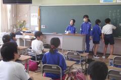 教室の教卓に立つ2人と黒板に文字を書く2人を見ている子供たちの写真