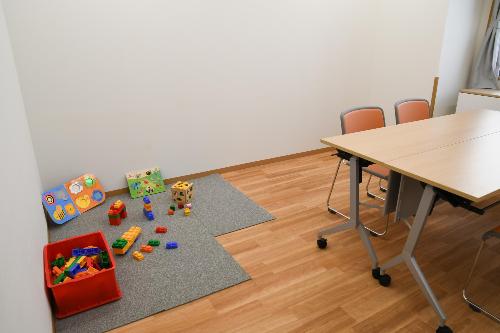 茶色の長テーブルとその周りに2つのオレンジ色の椅子、床にこども用のおもちゃが置かれている相談室の写真