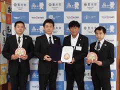 スーツ姿の男性4名が、それぞれ協定書と豊川市高齢者みまもり隊ワッペンを両手に持ち横に並んで立っている写真