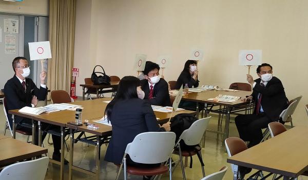 室内に組まれた長机を囲んで座り、丸印が描かれた手札を一斉に掲げて示している数人のスーツ姿の男女のグループの写真
