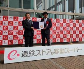 「遠鉄ストアの移動スーパー」と書かれた白い看板の設置された壇上で、スーツ姿の男性2名が協定書を料亭に持ち笑顔で立っている写真