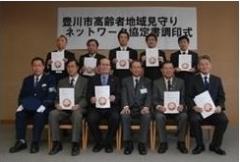 「豊川市高齢者地域見守りネットワーク協定書調印式」と書かれた白い幕の前にスーツ姿の男性が前列は椅子に座り、後列は立って横に並んでいる様子の写真