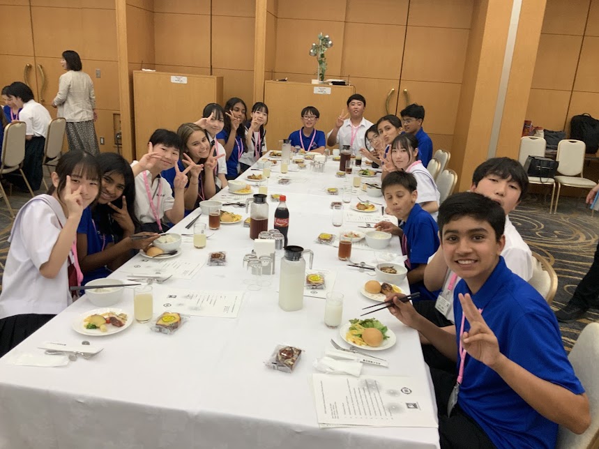 青い服を着たキュパティーノの中学生と制服を着た日本の中学生が食事を前にピースしている様子