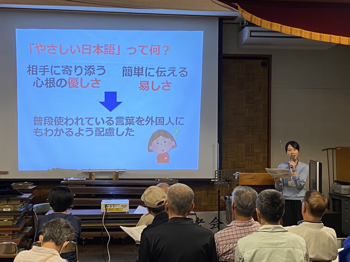 椅子に座った人たちが緑色のパイプ椅子に座ってスクリーンに映ったパワーポイント画像を見ている写真
