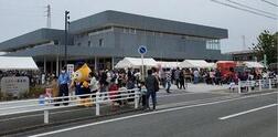 道路横の歩道で奥にイベントテントが立っておりたくさんの人が集まっている写真