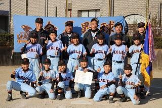 茶色い建物の前に3列に並んで整列している青い野球のユニフォームを着た子供達と黒い服を着た男性の写真