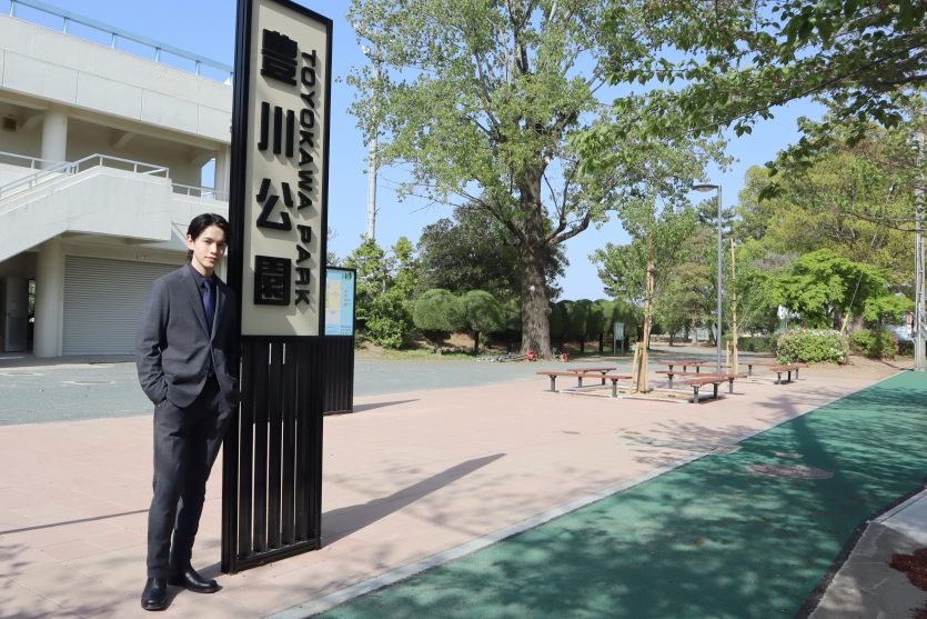 「豊川公園」書かれた看板の横にスーツ姿で立っている男性の写真
