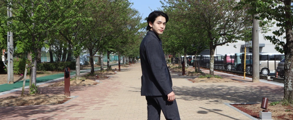 左右に木々が植えられた歩道のような所にスーツを着た男性が振り返っている写真