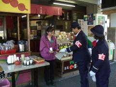 左腕に腕章をつけ制服を着た2人のクラブ員が店先で店員に話しかけている場面の写真
