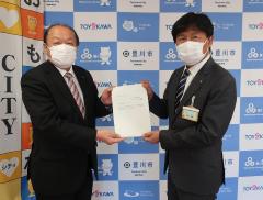 豊川市のバックパネルの前で2人のスーツとマスク姿の男性がそれぞれの手で1つの書類を持ち合いながらこちらに顔を向けている写真