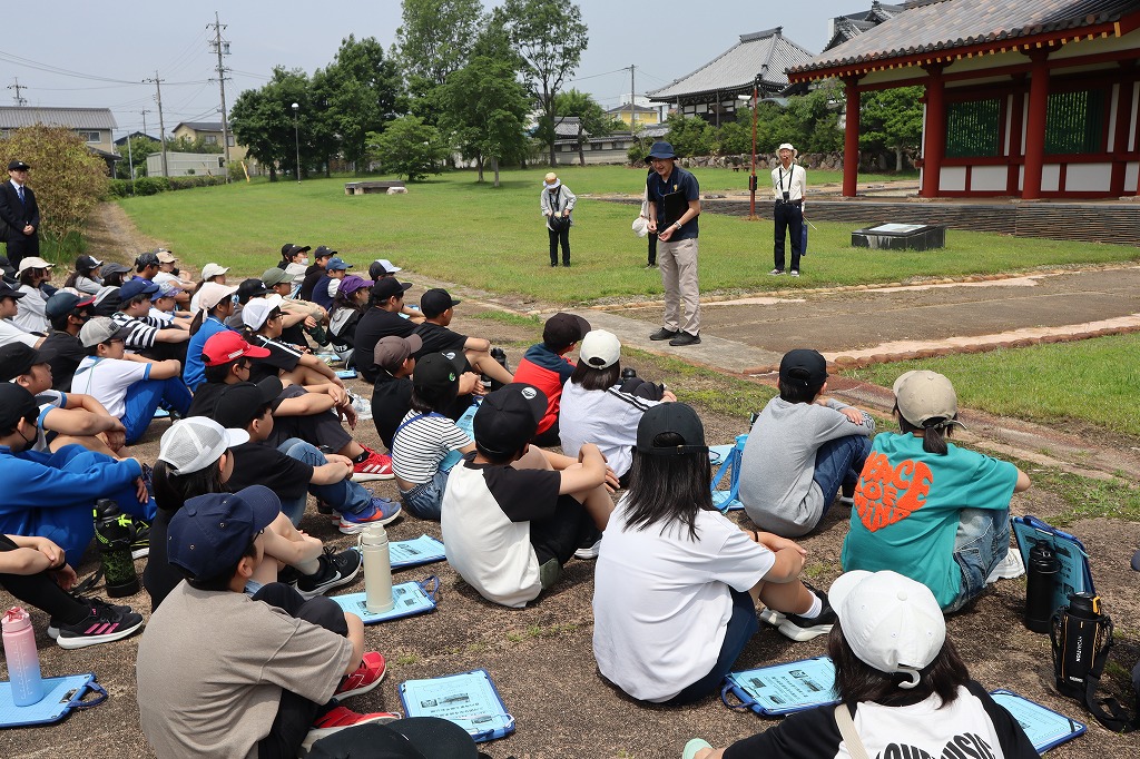 三河国分寺跡史跡公園で学年全体でガイドさんの話を聞く写真