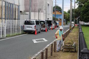 赤い5つのパイロンのそばに5台のワゴン車が駐停止している道路の右脇の雑草が散乱している歩道に置かれた刈り取った雑草が積まれた台車のそばに立つ黄色いの半袖の服を着て白い帽子を被り肩に水色のタオルをかけた男性の写真