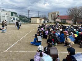 ランドセルを背負った児童たちが校庭にしゃがみ前に立つ児童の話を聞いている写真