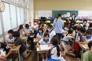 教室で机に座っている生徒たちと立っている女子児童と女性教師の写真