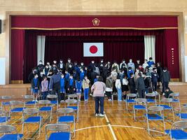 児童たちが体育館のステージの前で整列し先生が声掛けをしている写真