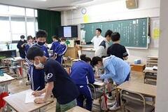 教室で席を立ち用紙を見ている児童たちと、黒板の前に立っている大人の写真