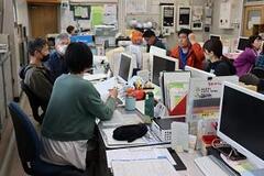 パソコンの並ぶ席に着席し、会話をする教師達の写真