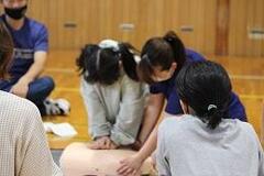 室内の床に座って心肺蘇生の訓練で使われるマネキンに心肺蘇生を行う人と、それを支える人の写真