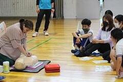 室内の床に座って心肺蘇生の訓練で使われるマネキンに心肺蘇生いている大人と、それを眺めている数人の人々の写真
