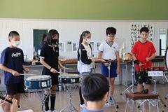 学校の音楽室で5人の児童がドラムなどの楽器の演奏をしており、その反対側で席に着いた児童たちがリコーダーを吹いている写真