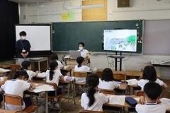 学校の教室で黒板の前でパソコンをしている先生と机に座っている児童たちの写真