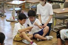 学校の教室で児童3人が床に座りながら端の1人の手元を見ている写真