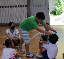 体育館で4人の子供たちが体育座りで座っており一人の男の子が1人の子の首にひも上のものをかけている写真
