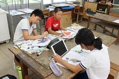 作業用テーブルに3人の子どもが座って用紙に絵をかいたりタブレットを見ている写真