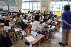 教室の席に座っている児童たちと、その間に座る大人たちと、後ろ手を組んで児童の方に向き合って立っている大人の写真