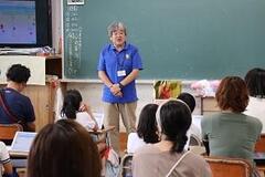 教室の黒板の前で手を前に組んで立っている一人の大人と、そちらを向いて座っている児童や大人たちの写真